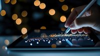 Hand drawing on digital tablet screen with stylus pen against blurred golden bokeh lights creating magical sparkle trail effect in dark background.