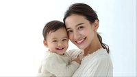 Young Asian mother in white sweater holding laughing baby in cream colored outfit against white background, expressing joy and tender family connection.
