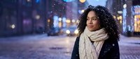Young African American woman with curly hair wearing knit scarf enjoying snowy evening in urban city street with bokeh lights and falling snowflakes.
