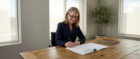 Professional blonde businesswoman in navy suit signing documents at wooden desk in modern office with natural lighting and plant decor.