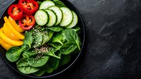 Fresh colorful vegetables and spinach leaves on black plate, sliced cucumber, yellow bell pepper, cherry tomatoes with sesame seeds, healthy food styling.