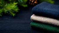 Folded natural linen fabrics in navy blue, beige and forest green colors on dark wooden background with pine branches and cone, sustainable textile concept.