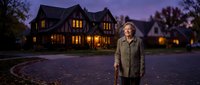 Senior woman standing confidently outside luxury home at twilight with warm interior lighting creating welcoming atmosphere for real estate concepts.