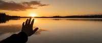 Hand reaching toward golden sunset over calm lake water with silhouetted trees and peaceful reflection for meditation and spiritual wellness concepts.