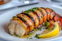 Grilled lobster tail sliced and seasoned with herbs, butter sauce, and fresh lemon wedge served on white plate, close up view of gourmet seafood dish.