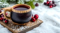 Hot winter beverage in black ceramic mug with wooden rim on rustic board, decorated with red Christmas ornaments and cranberries against snowy background.