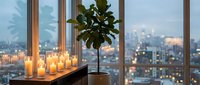 Cozy evening apartment interior with candles and fiddle leaf fig plant by large windows overlooking illuminated city skyline at dusk creating warm ambiance.