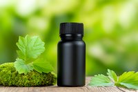 Black essential oil bottle with fresh green leaves on moss-covered surface against blurred natural background for aromatherapy and wellness products.