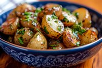 Roasted baby potatoes garnished with fresh herbs and garlic in decorative blue ceramic bowl, close up view of homemade side dish on wooden table.