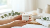 Hands holding white cosmetic jar with skincare product, displayed on wooden surface with fresh eucalyptus leaves and towels, ideal for beauty and wellness marketing.