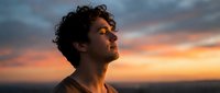 Young man with curly hair looking up peacefully at colorful sunset sky, expressing hope, gratitude and spiritual connection during golden hour meditation.