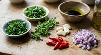 Fresh herbs and vegetables prepared for cooking on wooden cutting board with olive oil and balsamic vinegar for healthy Mediterranean cuisine preparation.