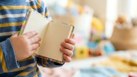 Child in striped shirt holding open book with blurred playroom background, educational reading concept for learning and literacy development.
