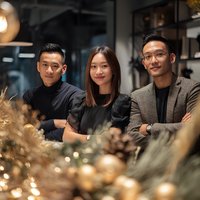 Three young Asian business professionals smiling together in modern office space with warm lighting and contemporary interior design elements.