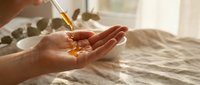 Woman applying golden facial serum oil from dropper bottle onto palm in bright natural sunlight for skincare routine and beauty treatment.