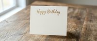 Elegant cream birthday card with gold lettering on rustic wooden table surface for celebration greetings and party invitations.