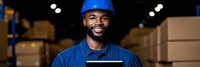 Smiling African American warehouse worker in blue uniform and hard hat standing confidently in industrial storage facility with cardboard boxes.