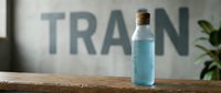 Young woman with light skin tone, professional appearance, holding a clear glass bottle containing blue liquid, positioned on wooden surface with blurred "TRAIN" text background and green plant, suitable for wellness, hydration, fitness, and supplement marketing materials.