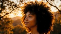 Young African American woman with natural curly afro hair enjoying peaceful moment outdoors during golden hour sunset with warm bokeh light background.