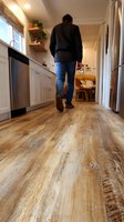 Person walking through modern kitchen with rustic wooden flooring, white cabinetry, and dining area visible in background.