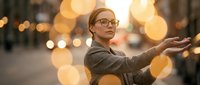 Young Caucasian woman with glasses poses gracefully on a city street at golden hour, surrounded by glowing bokeh lights, evoking confidence and urban elegance.