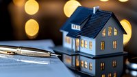 Miniature blue house model with glowing windows against blurred golden bokeh lights, silver pen on reflective surface. Real estate contract concept.