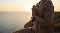 Woman in beige robe practicing meditation with prayer hands at golden hour sunset by ocean cliff for mindfulness and spiritual wellness retreat.