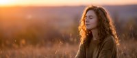 Young woman with curly hair enjoying peaceful sunset moment in golden field, eyes closed in meditation and tranquility outdoors.