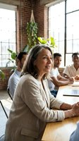 Mature woman with long gray hair and light skin tone, wearing a beige blazer, smiles while leading a business meeting in a modern loft office with brick walls and large windows. Use for corporate training, leadership, diversity, and workplace collaboration content.