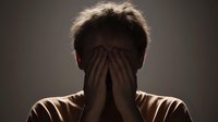 Silhouette of person covering face with hands in dark room, dramatic backlighting creates moody atmosphere expressing grief, depression, anxiety or emotional distress.