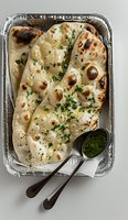 Freshly baked garlic naan bread topped with herbs and served in aluminum container with green chutney sauce, traditional Indian flatbread for restaurant menu.