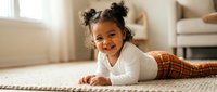 Happy African American toddler girl with curly hair in space buns lying on living room rug wearing white top and plaid pants smiling at camera in cozy home interior.