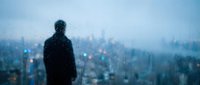 Silhouette of man standing by window with raindrops overlooking blurred city lights at dusk. Contemplative mood for mental health and solitude concepts.
