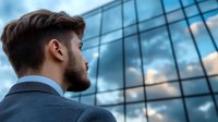 Profile view of young Caucasian businessman looking up at modern glass office building exterior reflecting dramatic cloudy sky, representing vision and aspirations in corporate world.