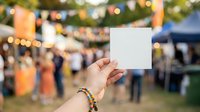 Hand holding blank white card at outdoor festival or event with blurred crowd and decorative lights in background, suitable for mockup, advertising, and promotional designs.