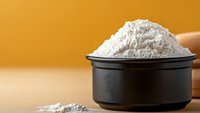 White flour in black ceramic bowl against warm orange background, with scattered flour powder on wooden surface, minimalist food photography setup.
