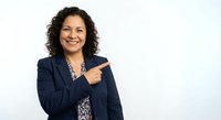 Professional Hispanic businesswoman in navy suit pointing finger gesture smiling confidently on white background for corporate presentations and marketing.