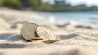 Golden coins lying on sandy beach with blurred tropical ocean and coastline in background, warm sunlight creating soft shadows on sand surface.