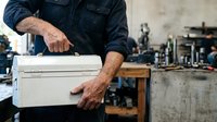 Mature male worker with tattooed arms holding white toolbox in industrial workshop setting. Skilled tradesman in dark work shirt carrying equipment in garage.