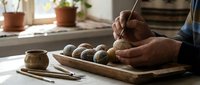 Potter sculpting clay balls on wooden board in sunlit studio workspace with tools and plants creating handmade ceramics for artistic pottery projects.