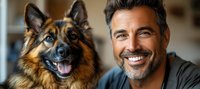 Mature man with stubble smiling next to German Shepherd dog, both looking at camera in warm indoor setting.