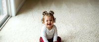 Happy toddler girl with curly hair sitting on carpet floor by window, smiling joyfully in bright natural light for family lifestyle photography.