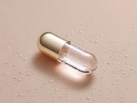 Clear transparent capsule with silver end on wet surface with water droplets, macro photography showing pharmaceutical detail in soft pink lighting.