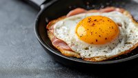 Sunny side up fried egg with crispy bacon in cast iron pan on dark background, seasoned with black pepper, close up view with copy space for breakfast menu.
