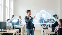 Young Caucasian businessman presenting cloud computing technology in modern office environment with colleagues working at desks in background.
