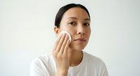 Asian woman applying skincare product with cotton pad on face during daily beauty routine against white background for cosmetic advertising.