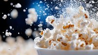 Popcorn kernels exploding with dramatic splash effect against dark background, crisp white popped corn in motion with bokeh lights and flying particles.