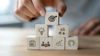 Hand arranging wooden blocks with business strategy icons including target, lightbulb, team, chart, and innovation symbols on office desk.
