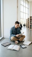 Young Caucasian male student sitting cross-legged on library floor reading book surrounded by tablets and open books near large windows with natural light.