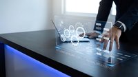 Businessman in dark suit interacting with holographic gears and digital interface on touchscreen table, representing technology innovation and business solutions.